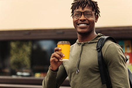 Image of smiling hispanic man, with glasses walking down city street holding takeaway coffee, happy man enjoying hot drink outdoorsの写真素材