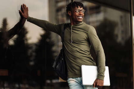 Curly American business man, with a backpack, holding a laptop, posing with glasses in front of the...