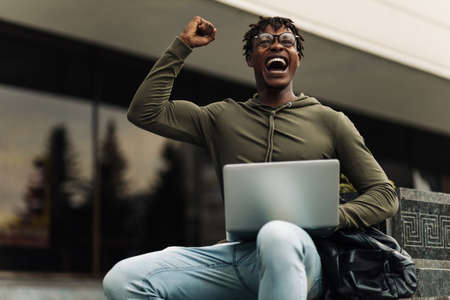 Excited African man, celebrating success, reading good news by email, businessman looking at laptop, showing yes gesture and laughing while sitting outdoors in city against glass building backgroundの写真素材