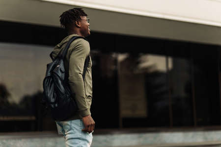 Happy handsome successful african student, wearing glasses and with a black backpack, standing on the steps and smiling, looks into the frame outdoors on the street near the universityの写真素材
