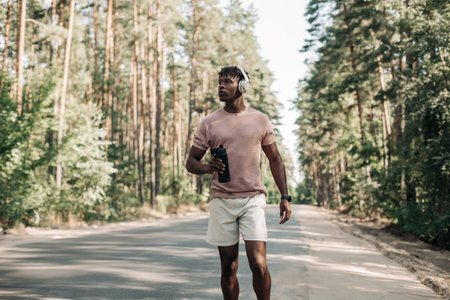 Sporty tired african american man, runner drinking water resting after workout, man relaxing after workout on city road in forest in natureの写真素材