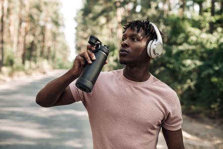 Sporty tired african american man, runner drinking water resting after workout, man relaxing after...
