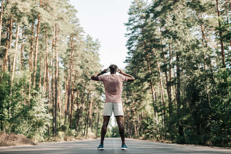 Black sporty man, dressed in sportswear listening to music on headphones during morning workout,...