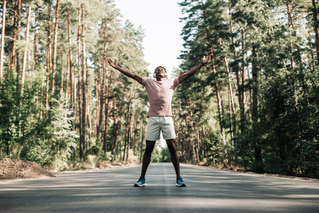 African American fitness man, in sportswear and in headphones, standing on the road in the forest at dawn, with his arms raised, enjoying freedom, Winnerの写真素材