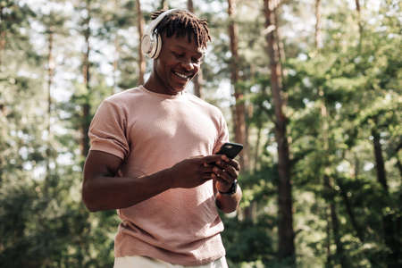 Happy smiling black sportsman, listening to music on headphones and using phone while relaxing, exercising in a park outdoorsの写真素材