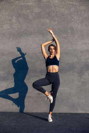 Beautiful smiling happy young woman, in sports fitness clothes, workout against a gray wall, doing...