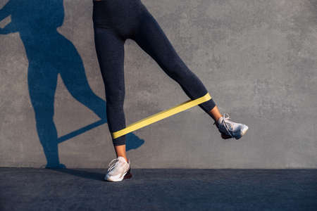 Beautiful sporty woman doing exercises with resistance band, fitness woman doing leg exercises with yellow fitness rubber band on gray backgroundの写真素材