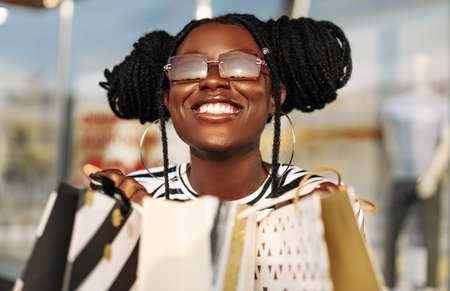 Happy fashionable African American woman in sunglasses, with shopping bags looking at a shop window in the city, shopping for Black Friday on saleの写真素材