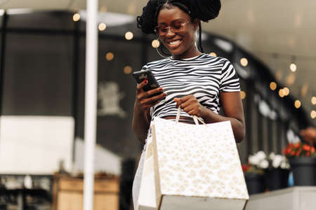 Beautiful fashionable african american girl holding bags, using smartphone and smiling while shopping at womens boutique for Black Friday, shopping concept, black friday saleの写真素材