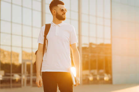 Young stylish man with a beard in sunglasses, with a briefcase and a laptop in his hands. A man in the city against the backdrop of a glass building in the background. A man on the street at sunsetの写真素材