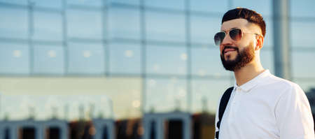 handsome man with a beard in sunglasses, in a casual white shirt on a sunny day, smiles. Man outdoors near a glass building, Fashion man, on a street backgroundの写真素材