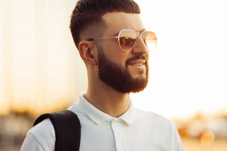 handsome man with a beard in sunglasses, in a casual white shirt on a sunny day, smiles. Man outdoors near a glass building, Fashion man, on a street backgroundの写真素材