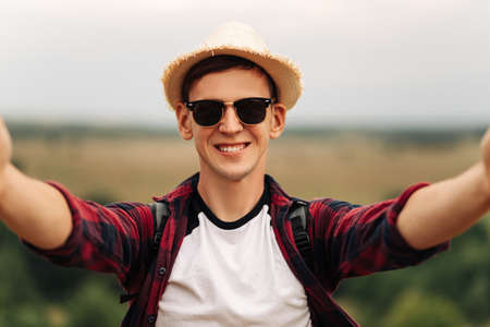 Young male tourist in sunglasses and a hat, makes a selfie while walking in the woods. A traveler leading a healthy active lifestyle, enjoying nature, walking in nature, summer walk in the mountainsの写真素材