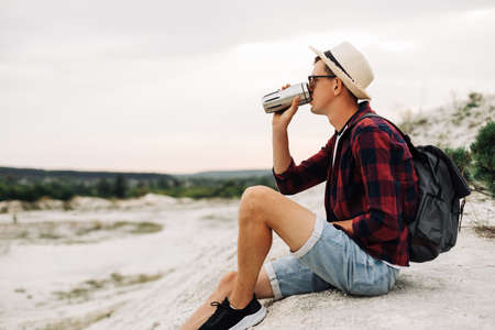 hiker with a backpack sits on the top of the mountain, enjoying the view of the coast. A young man with a cup of coffee sits on a rock with a view of the nature around. Mountain and coast travelの写真素材