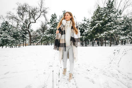 Young beautiful woman in winter warm clothes, in a sweater and a scarf walks through the forest in...