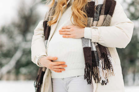 pregnant woman in warm winter clothes standing outside on a snowy winter day, with hands on her stomach on the background of nature. Concept of pregnancy, motherhood, expecting the birth of a childの写真素材