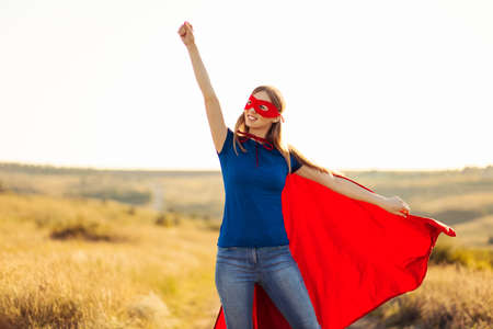 superhero women. A young and beautiful blonde in the form of a superheroine in a red cloak is growing Forward and upward. Woman dressed as a superhero in nature. Feminism conceptの写真素材