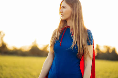 Joyful beautiful woman in a superhero costume posing against the backdrop of a sunset in nature. superhero women. Young and beautiful blonde in the image of a superheroine with a red cloakの写真素材