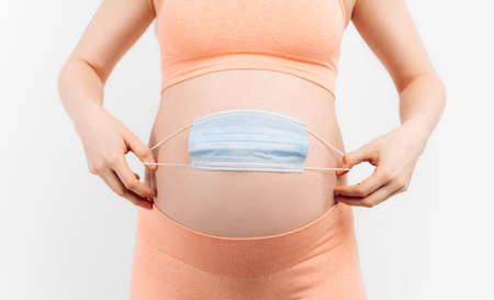 Belly of a pregnant woman holding a medical mask close-up on a white background. Pregnancy of a woman and a medical mask on her stomach, the concept of protection from epidemicsの写真素材