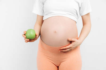 Close-up of a young pregnant woman holding a green apple on a white background. Expectant mother is...