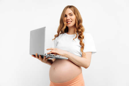 Happy pregnant woman using laptop, browsing internet and reading pregnancy blogs, isolated on white background. Pregnant woman working on laptop at homeの写真素材