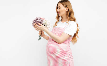 Beautiful pregnant woman with flowers holds her hands on her stomach on a white background. A young woman is waiting for the birth of a child. Pregnancy, motherhood, mother's day holiday conceptの写真素材