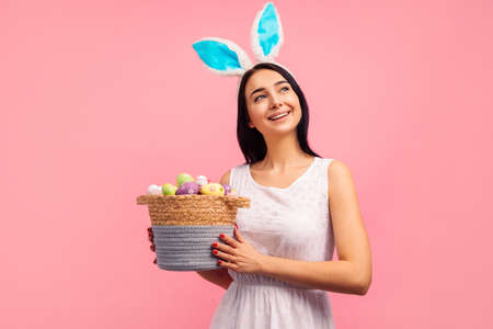 Happy woman in rabbit ears holding a basket with colored eggs, a traditional holiday, in the studio...