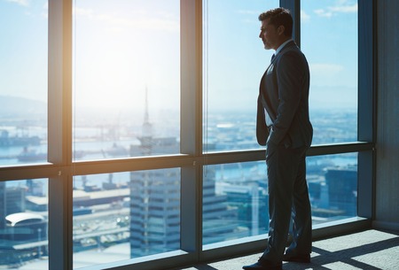 Mature business executive and leader standing confidently with hands in his pockets, in the the top floor office, looking at the city below through large windowsの写真素材