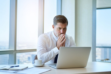 Handsome and mature business executive sitting at his desk in amodern office with large windows looking seriously at information on the screen of his laptop computer.の写真素材