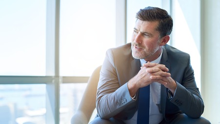 Mature businessman with a handsome and modern look, sitting in an office space looking away thoughtfully with a positive expressionの写真素材