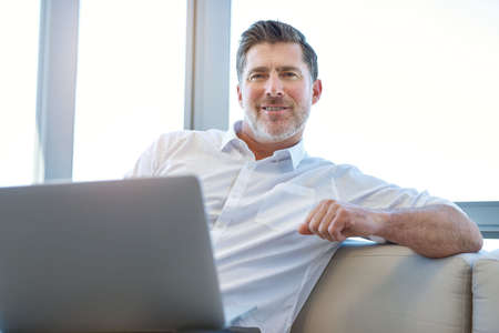 Portrait of a mature businessman sitting comfortably with a laptop computer on his lap, smiling...