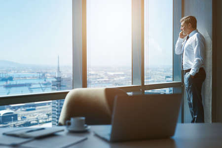 Handsome mature businessman standing next to the large windows of his top floor office, looking at...