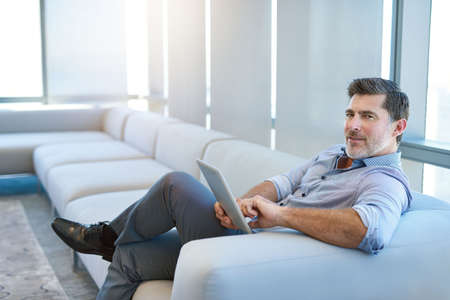 Portrait of a handsome mature businessman, relaxing on a couch in a business lounge, using a digital...