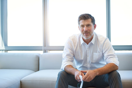Handsome mature businessman sitting on a couch with large windows behind him, holding some paperwork...