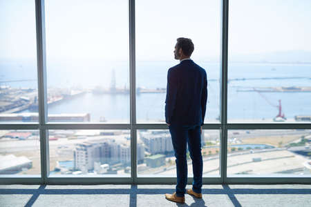 Full length rearview of a young stylish businessman standing at the large windows of a high office, looking at the view of a harbour belowの写真素材
