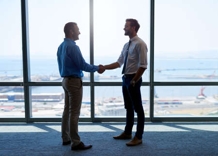 Full length shot of two businessmen shaking hands to acknowledge their partnership, and smiling positively, with a large office window behind themの写真素材