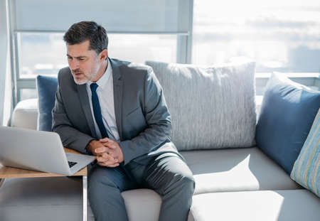 Mature businessman in a suit sitting on a sofa in a modern office using a laptopの写真素材