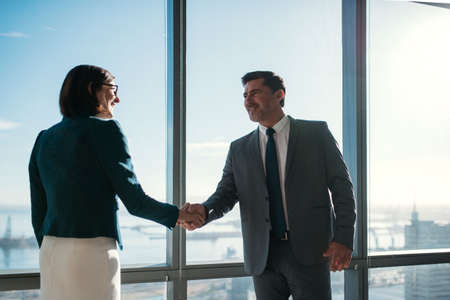 Businessman and businesswoman shaking hands together while standing in front of office building windows overlooking the cityの写真素材