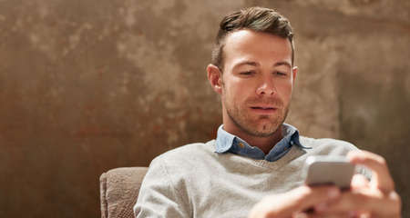 Casually dressed young businessman reading a text message on a cellphone while sitting in a modern...