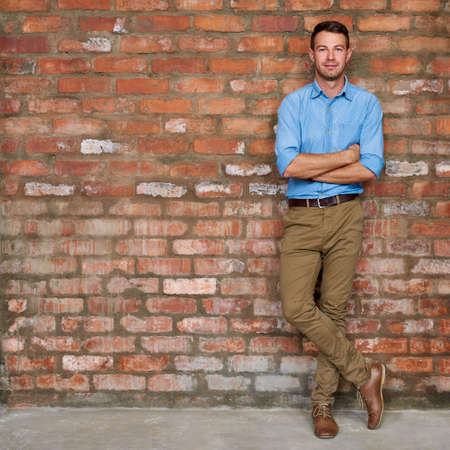 Portrait of a casually dressed young businessman standing with his arms crossed against a brick wall in an officeの写真素材