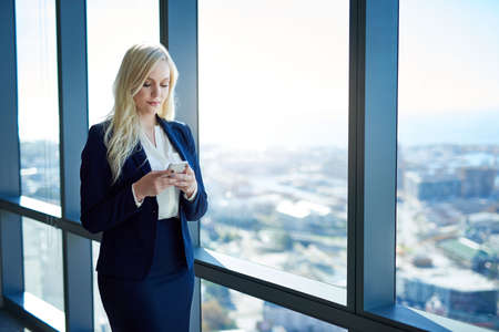 Young businesswoman reading a text message on her cellphone while standing by windows in a modern officeの写真素材