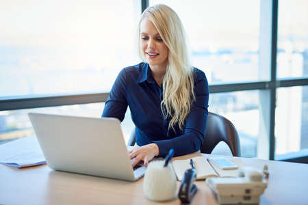Smiling young businesswoman sitting alone at a desk in a bright modern office working on a laptopの写真素材