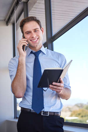 Portrait of a friendly young businessman standing in his office with his diary open in his hand,...