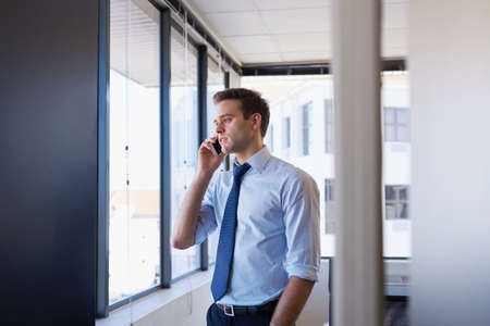 Handsome young employee standing in his business office talking on his mobile phone while looking out of the windowの写真素材