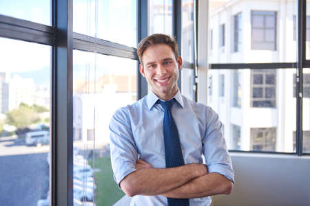 Friendly young employee of a corporate business, standing proudly in his corner office in front of large windows, smiling confidently at the camera with his arms crossedの写真素材