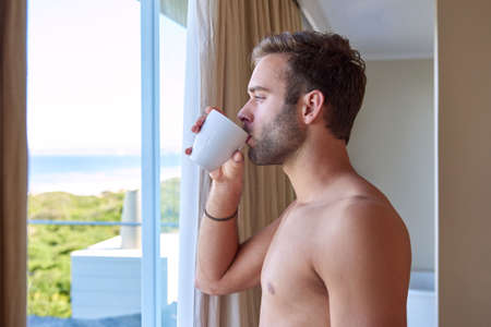 Handsome young man standing inside looking at a view of the beach through his hotel window, while sipping his first cup of coffee for the morningの写真素材