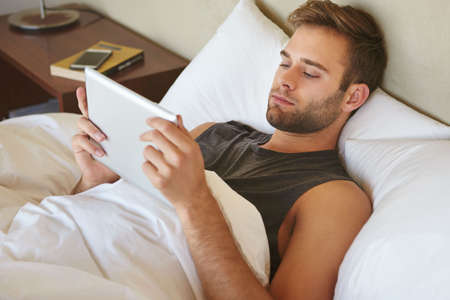 Handsome young man with stubble, relaxing in bed at home and reading an ebook on his digital tablet,...