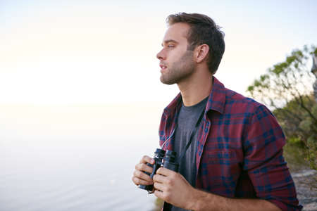 Handsome young male student looking out to the horizon while holding binoculars outdoors in nature