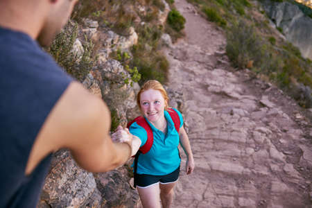 Athletic young woman with red hair holding the hand of a guy offering her assistance on a rocky nature trailの写真素材