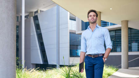 Confident young businessman standing with a digital tablet on a footpath outside of a modern office...